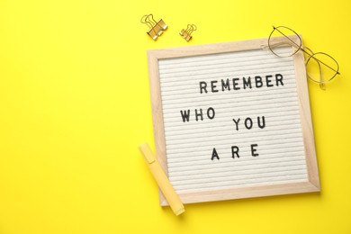 Photo of Letter board with phrase Remember Who You Are, glasses, marker and binder clips on yellow background, flat lay. Space for text