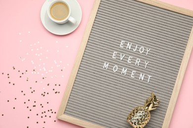 Photo of Letter board with phrase Enjoy Every Moment, cup of coffee, pineapple figure and confetti on pink table, flat lay