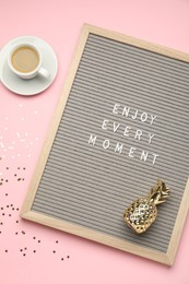 Photo of Letter board with phrase Enjoy Every Moment, cup of coffee, pineapple figure and confetti on pink table, flat lay