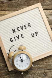 Letter board with phrase Never Give Up and alarm clock on wooden table, top view