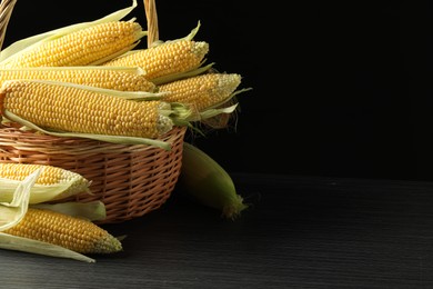 Many fresh ripe corncobs with green husks in wicker basket on table against black background, closeup. Space for text