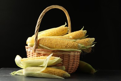 Many fresh ripe corncobs with green husks in wicker basket on table against black background