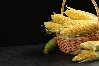 Photo of Many fresh ripe corncobs with green husks in wicker basket on table against black background, closeup. Space for text