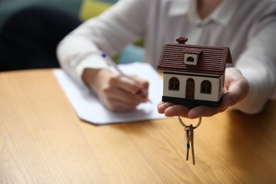 Photo of Woman working with document while holding house model and key at wooden table indoors, closeup