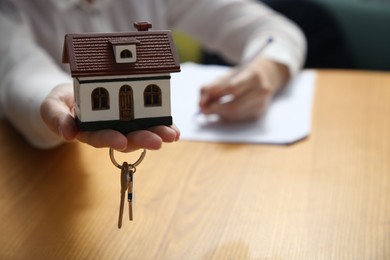 Photo of Woman working with document while holding house model and key at wooden table indoors, closeup