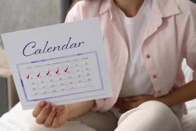 Photo of Woman holding calendar with marked menstrual cycle days on bed at home, closeup