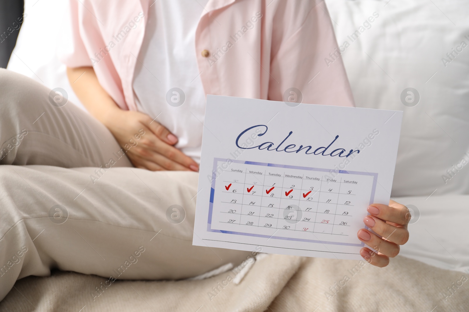 Photo of Woman holding calendar with marked menstrual cycle days on bed at home, closeup