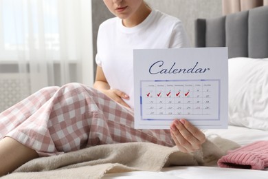 Photo of Woman holding calendar with marked menstrual cycle days on bed at home, closeup