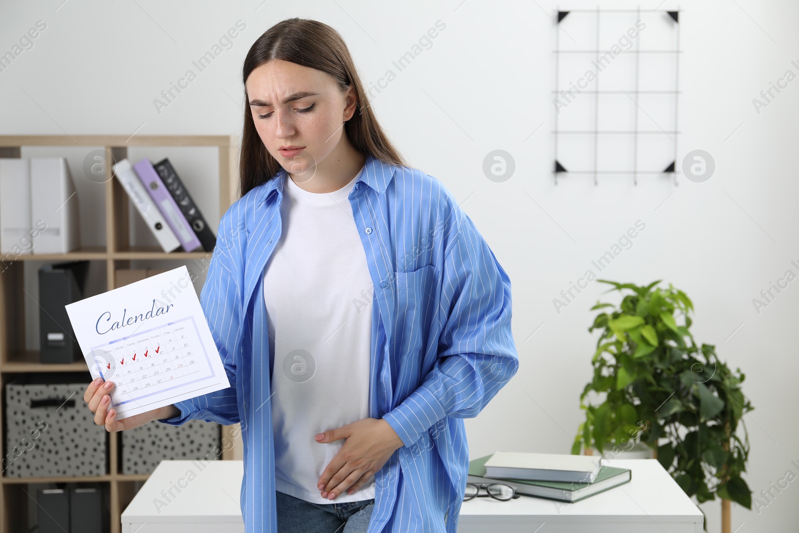 Photo of Woman holding calendar with marked menstrual cycle days and suffering from abdominal pain indoors