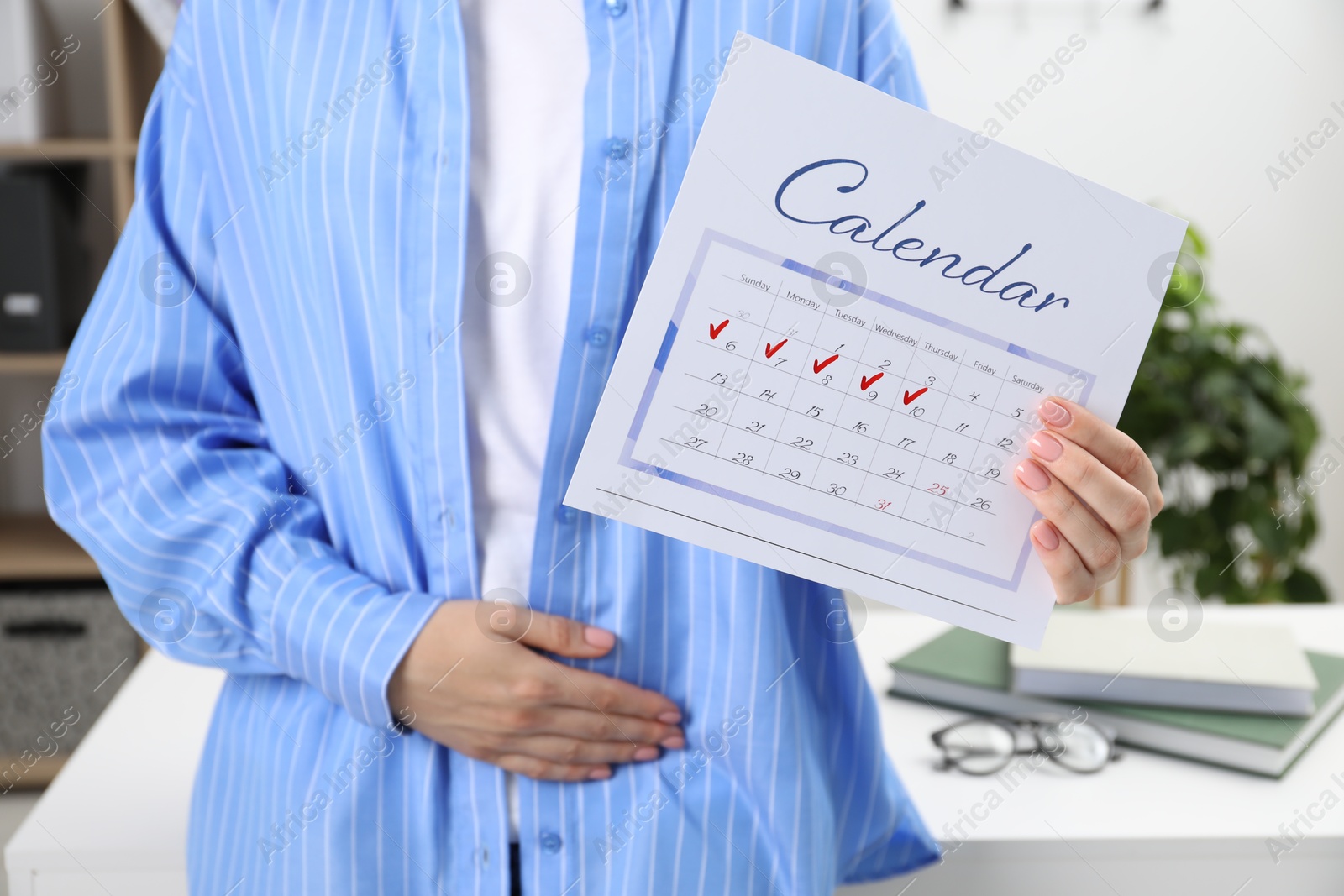 Photo of Woman holding calendar with marked menstrual cycle days and suffering from abdominal pain indoors, closeup