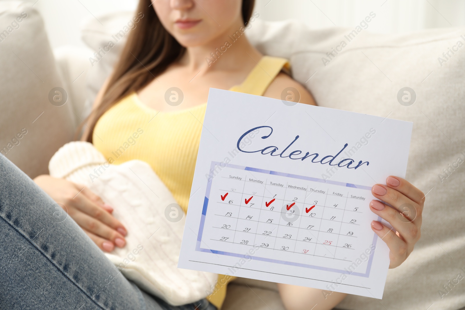 Photo of Woman holding calendar with marked menstrual cycle days and using hot water bottle on sofa at home, closeup