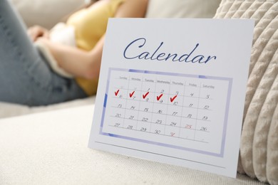 Photo of Woman using hot water bottle indoors, focus on calendar with marked menstrual cycle days on sofa