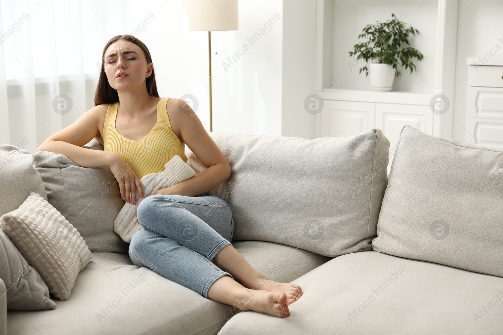 Photo of Woman using hot water bottle to relieve menstrual pain on sofa at home