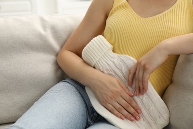 Photo of Woman using hot water bottle to relieve menstrual pain on sofa at home, closeup