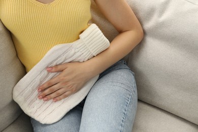 Photo of Woman using hot water bottle to relieve menstrual pain on sofa at home, closeup