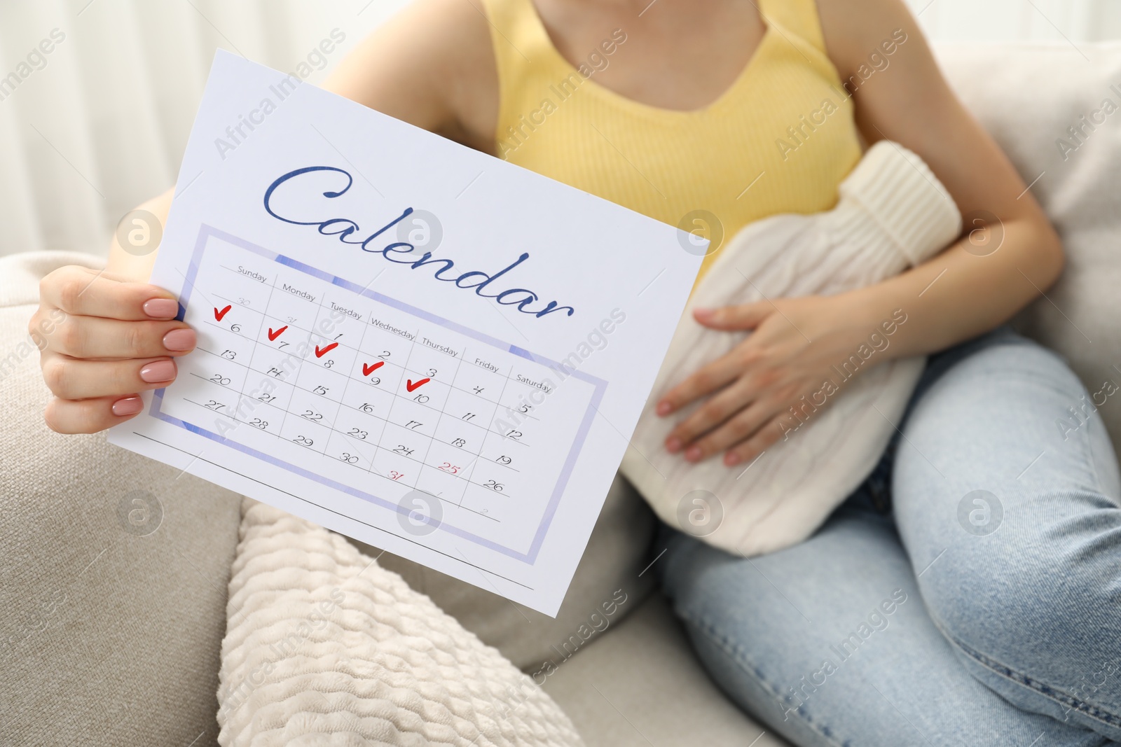 Photo of Woman holding calendar with marked menstrual cycle days and using hot water bottle on sofa at home, closeup