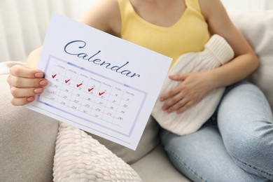 Woman holding calendar with marked menstrual cycle days and using hot water bottle on sofa at home, closeup