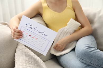 Photo of Woman holding calendar with marked menstrual cycle days and using hot water bottle on sofa at home, closeup