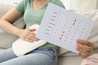 Woman holding calendar with marked menstrual cycle days and using hot water bottle on sofa at home, closeup