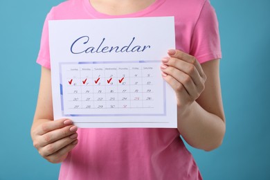 Woman holding calendar with marked menstrual cycle days on light blue background, closeup