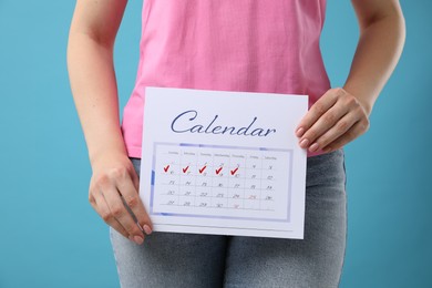 Woman holding calendar with marked menstrual cycle days on light blue background, closeup