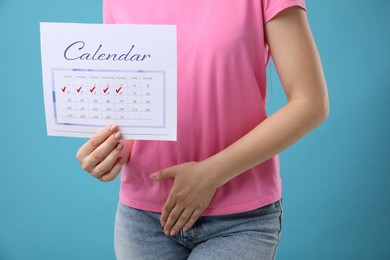 Woman holding calendar with marked menstrual cycle days and suffering from abdominal pain on light blue background, closeup