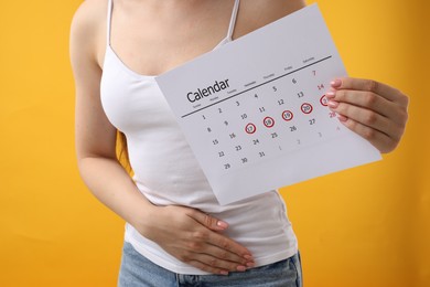 Photo of Woman holding calendar with marked menstrual cycle days and suffering from abdominal pain on orange background, closeup