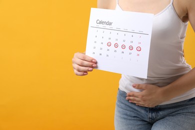 Woman holding calendar with marked menstrual cycle days and suffering from abdominal pain on orange background, closeup. Space for text