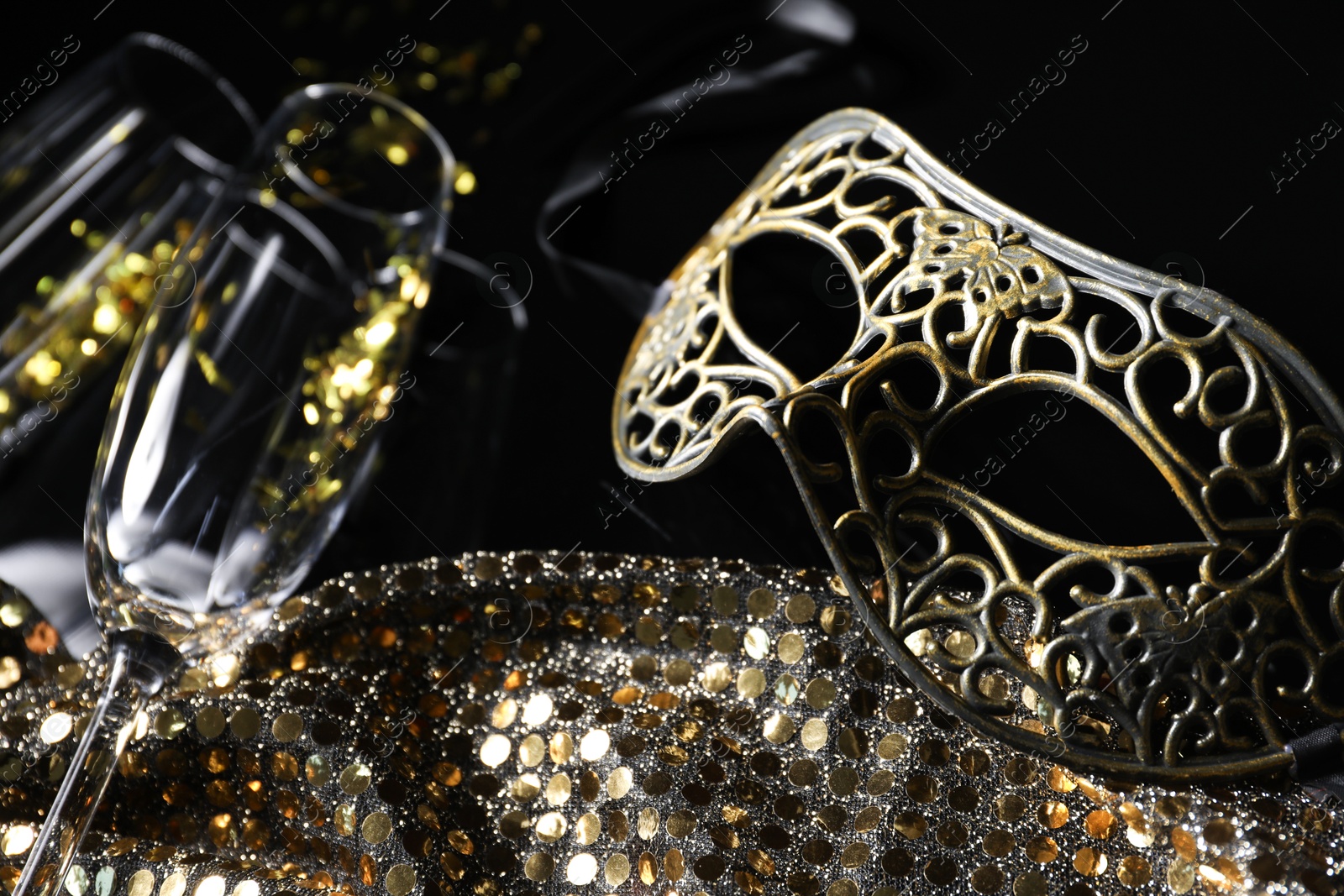 Photo of Beautiful carnival masks, champagne, glasses and fabric with golden sequins on black background, closeup