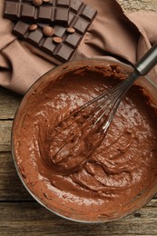 Photo of Chocolate dough in bowl and whisk on wooden table, top view