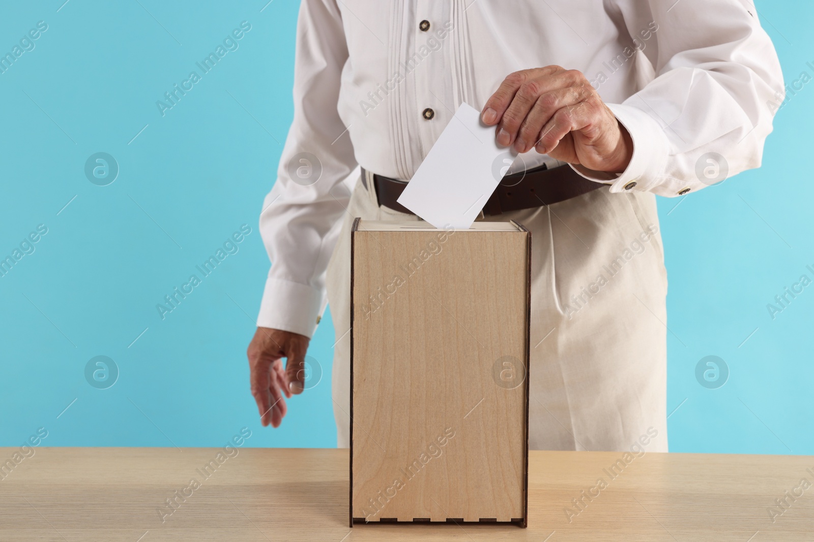 Photo of Referendum. Man putting his vote into ballot box at wooden table against light blue background, closeup