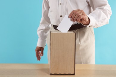 Photo of Referendum. Man putting his vote into ballot box at wooden table against light blue background, closeup