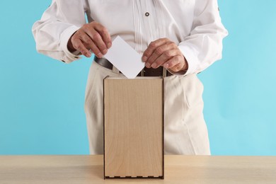 Photo of Referendum. Man putting his vote into ballot box at wooden table against light blue background, closeup