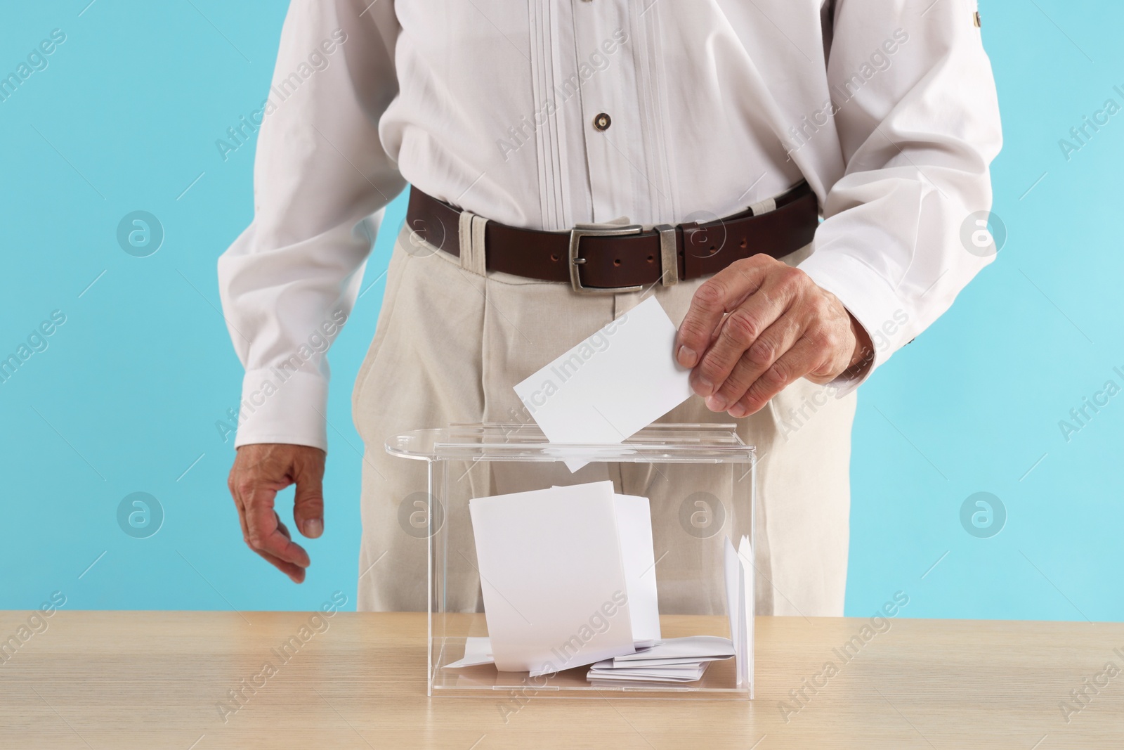 Photo of Referendum. Man putting his vote into ballot box at wooden table against light blue background, closeup