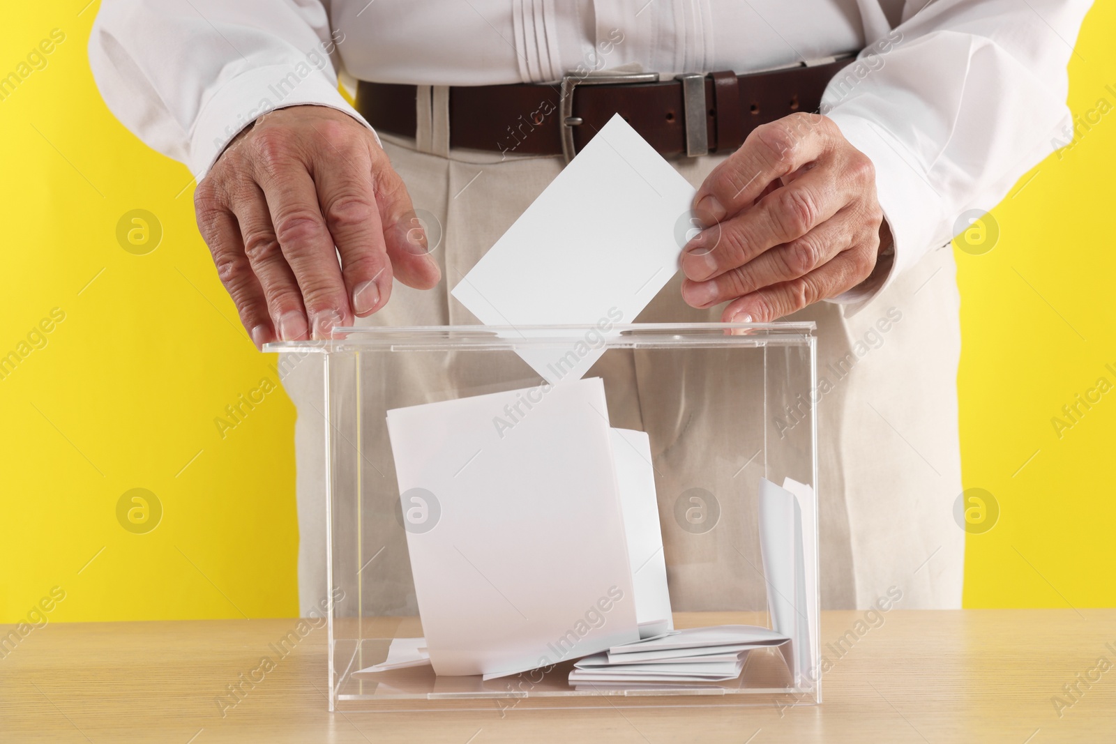 Photo of Referendum. Man putting his vote into ballot box at wooden table against yellow background, closeup