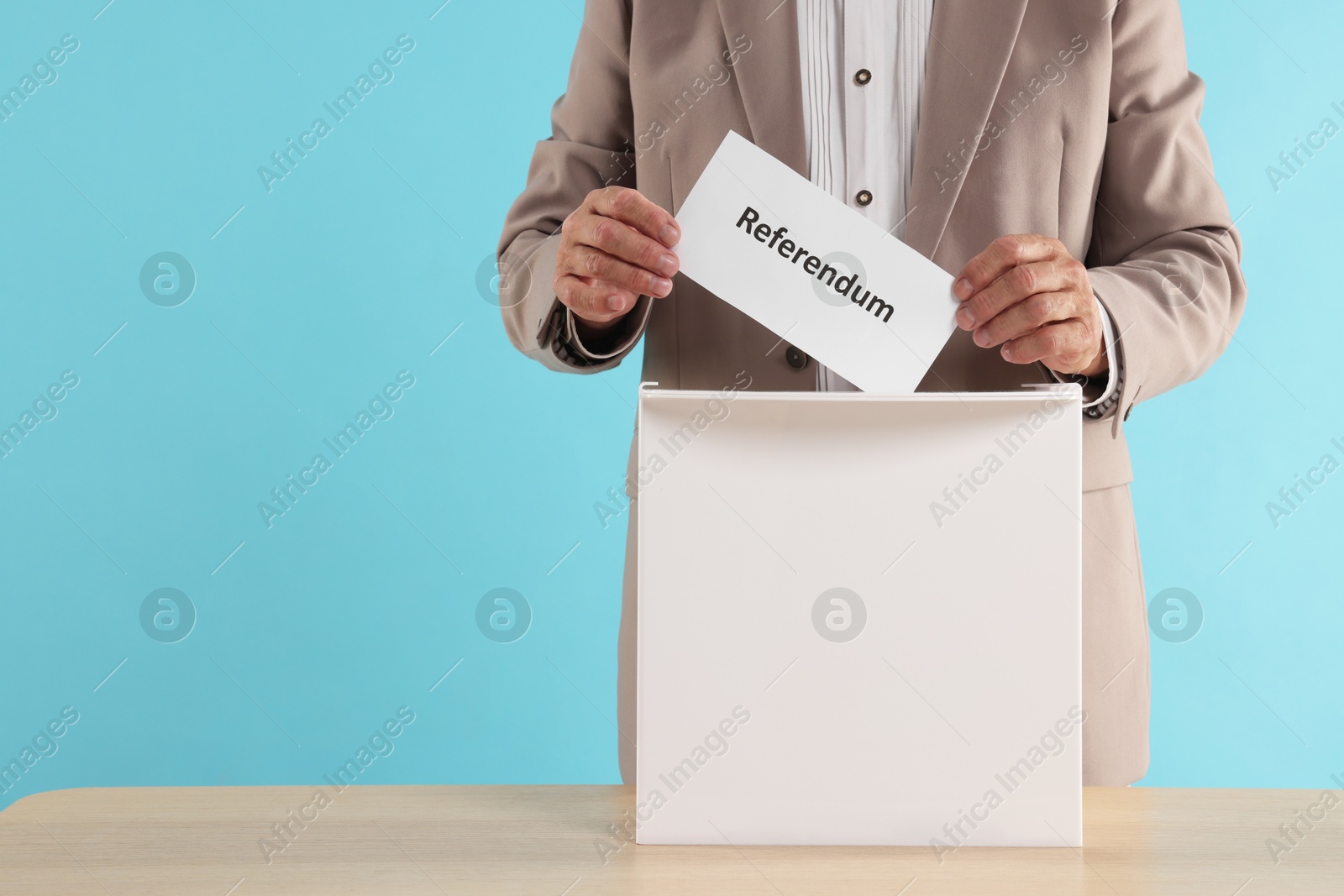 Photo of Referendum. Man putting his vote into ballot box at wooden table against light blue background, closeup