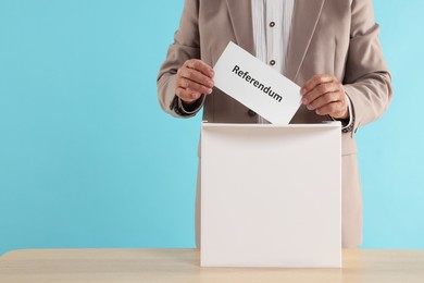 Referendum. Man putting his vote into ballot box at wooden table against light blue background, closeup