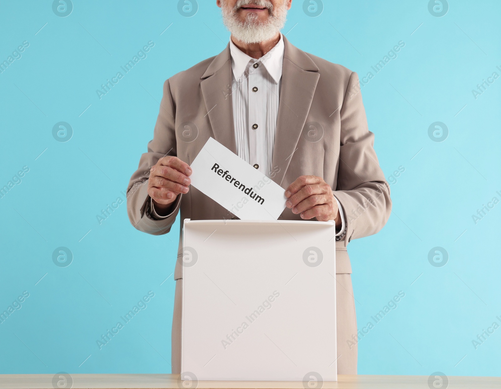 Photo of Referendum. Man putting his vote into ballot box at wooden table against light blue background, closeup