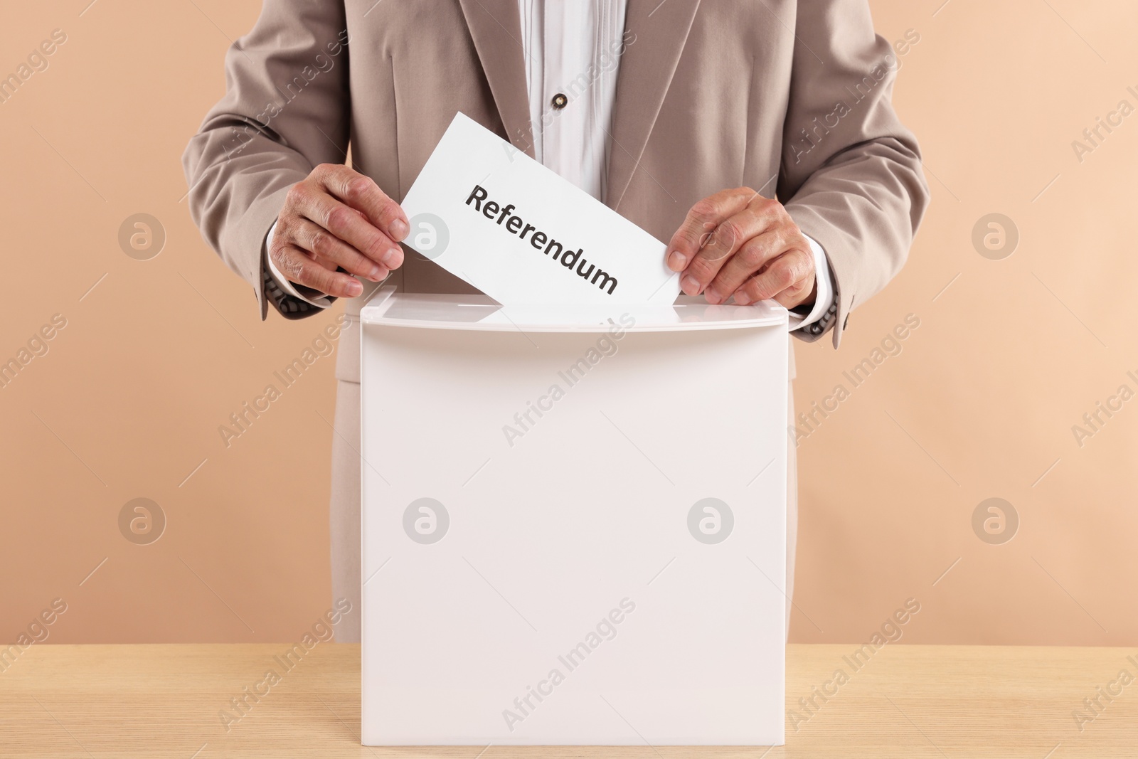 Photo of Referendum. Man putting his vote into ballot box at wooden table against beige background, closeup