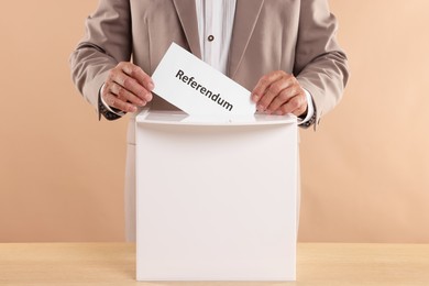 Referendum. Man putting his vote into ballot box at wooden table against beige background, closeup