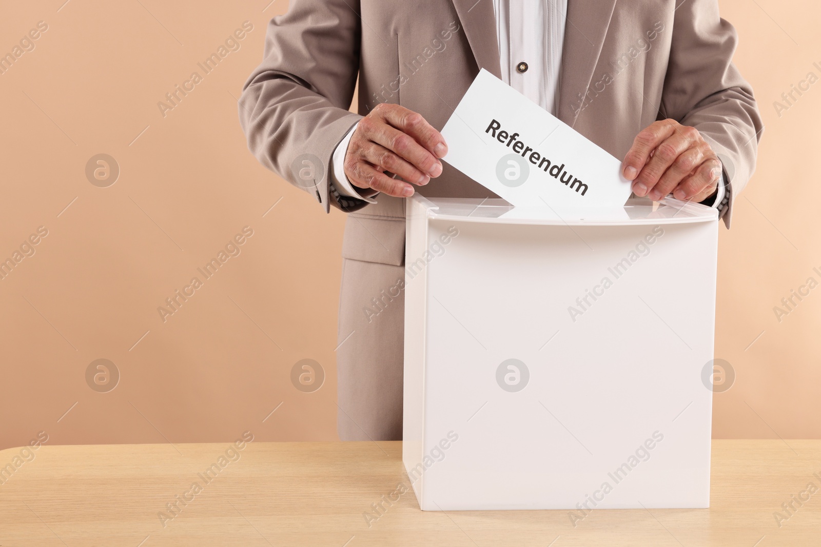 Photo of Referendum. Man putting his vote into ballot box at wooden table against beige background, closeup