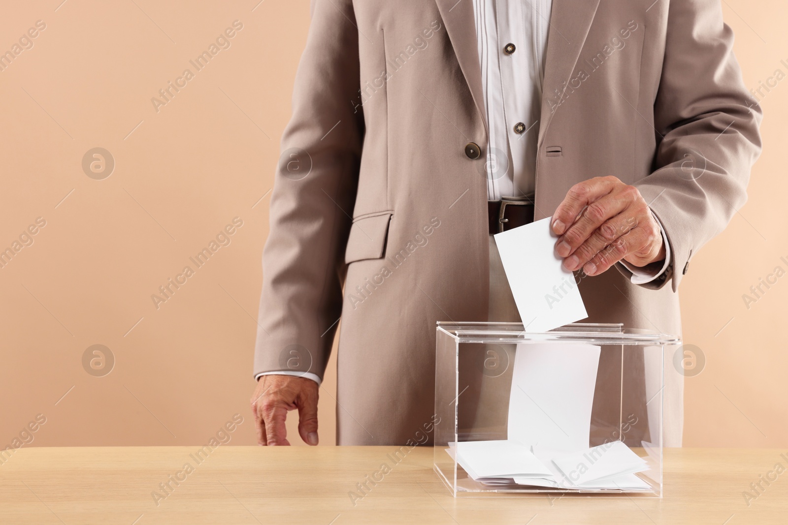 Photo of Referendum. Man putting his vote into ballot box at wooden table against beige background, closeup