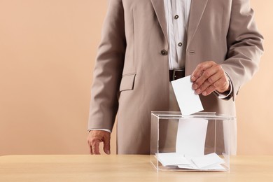 Referendum. Man putting his vote into ballot box at wooden table against beige background, closeup