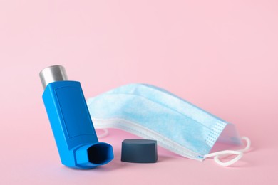 Photo of Asthma inhaler and medical mask on pink background, closeup