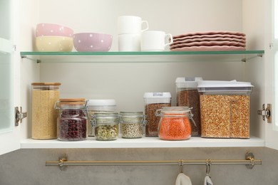 Photo of Different types of cereals, legumes in containers and tableware in closet