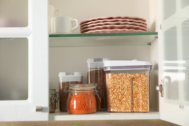 Photo of Different types of cereals, legumes in containers and tableware in closet