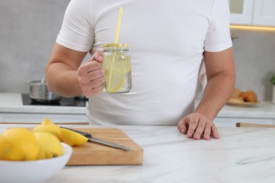 Photo of Man holding mason jar of water with lemon at white marble table in kitchen, closeup