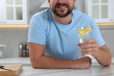 Happy man holding glass of water with lemon at white marble table in kitchen, closeup