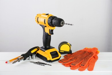 Photo of Cordless electric drill, gloves, bits, screwdriver and measuring tape on white wooden table