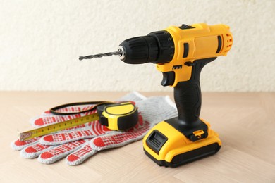 Cordless electric drill, gloves and measuring tape on wooden surface